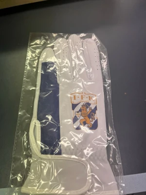 IFK Göteborg vit och blå handske - Snygg vit handske med blå detalj och IFK Göteborgs klubbmärke på ovansidan. Handsken har små ventilationshål och är tillverkad i ett slätt syntetmaterial. Perfekt för dig som vill visa klubbkänsla på träningen eller matchen.