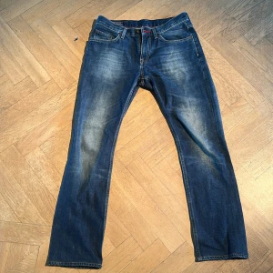 Tommy Hilfiger blå jeans 31/32 - Säljer ett par snygga blå jeans från Tommy Hilfiger i storlek 31/32. Klassisk femficksmodell med raka ben och diskret slitning framtill. Jeansen har röda sömmar på insidan och är tillverkade i robust denim. Perfekt för dig som gillar stilrena och tidlösa jeans.