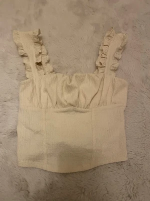 Beige linnetopp med volangaxelband - Supersöt beige linnetopp med breda axelband dekorerade med volanger. Toppen har en lätt rynkad struktur och är tillverkad i ett mjukt, krinklat material som känns luftigt. Perfekt för varma dagar och ger en romantisk vibe till din outfit.