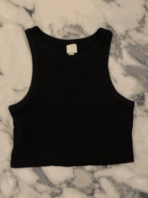 Svart croppad linnetopp från H&M - Svart croppad linnetopp från H&M i storlek S. Toppen har en klassisk rund halsringning och är ärmlös med breda axelband. Materialet är mjuk bomull som känns skönt mot huden. Perfekt att styla med jeans eller kjol för en chill look.