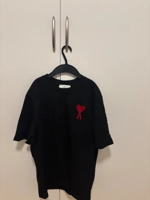 Svart t-shirt från AMI med rött hjärta - Svart t-shirt från AMI med klassisk rund hals och korta ärmar. På bröstet finns ett broderat rött hjärta med bokstaven A, vilket ger en snygg detalj. Materialet är mjuk bomull och passformen är loose. Perfekt för dig som gillar stilrena plagg med en twist.