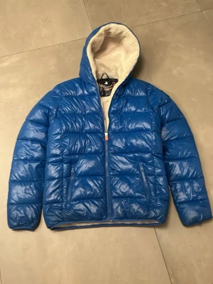 Blå pufferjacka med huva - Blå pufferjacka från Panda med huva och mjukt teddyfoder på insidan. Jackan har två fickor med dragkedja och quiltad design. Perfekt för kalla dagar när du vill hålla dig varm och ändå se fräsch ut. Liten defekt vid slutet av armen. Ny pris 3000kr