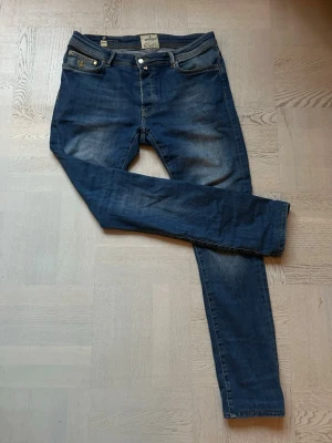 Blå jeans från Morris M 32/34 - Säljer ett par klassiska blå jeans från Morris M i storlek 32/34. Jeansen har snygga slitningar, guldfärgade detaljer. De har normal passform och raka ben, tillverkade i slitstarkt denimtyg.