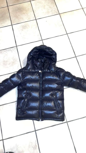  Mörk blå Moncler Maja  pufferjacka med huva - Säljer en mörklblå pufferjacka från Moncler Maja med glansig finish och huva.  Jackan har nfc och Scan. Jackan har två snedställda fickor med dragkedja, quiltad design och brunt innerfoder. Perfekt för kalla dagar och har en snygg, modern look.