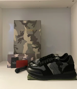 Valentino Rockrunner - Säljer ett par svarta och gråa Valentino Garavani sneakers med snyggt camouflage-mönster. Skorna har snörning, rund tå och detaljer i mocka och textil. Riktigt stilrena och unika sneakers som sticker ut med sin exklusiva design.