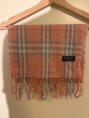 Burberry halsduk - Snygg rutig halsduk från Burberry i mjuk rosa, beige och grå lamull. Klassisk design med fransar nedtill och ikoniskt Burberry-märke. Perfekt för dig som vill ha en stilren och tidlös look med lite brittisk vibe. Skriv vid minsta fundering!🤩