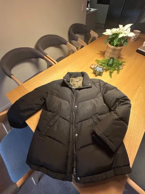 Svart pufferjacka från Tatum, stl 42 - Svart pufferjacka från Tatum i storlek 42. Jackan har en hög krage, dragkedja och tryckknappar framtill samt två snedställda fickor. Fodret är olivgrönt och jackan är perfekt för kalla dagar. Klassisk och enkel design som håller dig varm.