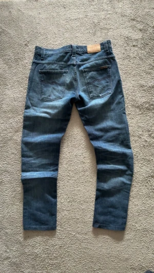 Blå jeans från Nudie Jeans - Säljer ett par klassiska blå jeans från Nudie Jeans med snygg tvätt och raka ben. Jeansen har fem fickor, synliga sömmar och läderpatch med logga bak i midjan. Perfekta för dig som gillar stilrena och tidlösa jeans.