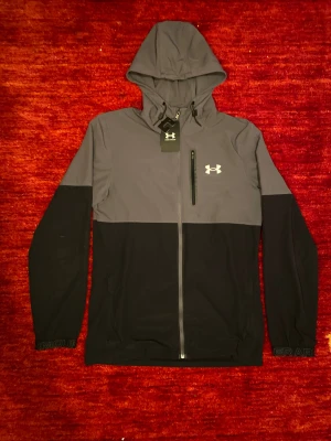 Grå och svart vindjacka Under Armour - Helt ny Under Armour-jacka i toppkvalitet, fortfarande med prislapp kvar. Lätt, bekväm och perfekt för både träning och vardag. Snygg tvåfärgad design med huva och vattentålig bröstficka. Aldrig använd – i absolut nyskick