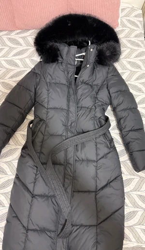 Svart lång pufferjacka med fuskpäls - Snygg svart pufferjacka med lång passform och huva med fluffig fuskpäls. Jackan har quiltad design, dragkedja och knappar framtill samt ett bälte i midjan för extra stilpoäng. Insidan är fodrad med mjuk pälsimitation som gör den extra varm under kalla dagar. Luvan går även att ta bort samt pälsen på luvan och pälsen på luvan är inte äkta ☺️