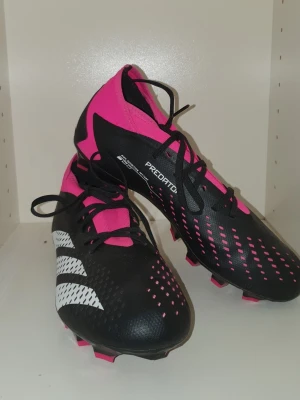 Adidas Predator fotbollsskor svart/rosa - Adidas Predator fotbollsskor i svart och rosa med vita detaljer. Skorna har snörning, platt sula med rosa dobbar och en snygg, sportig design. Tillverkade i syntetmaterial för bra grepp och komfort på planen. Perfekt för dig som vill sticka ut med färg och stil.kan gå ner till 300 kr 