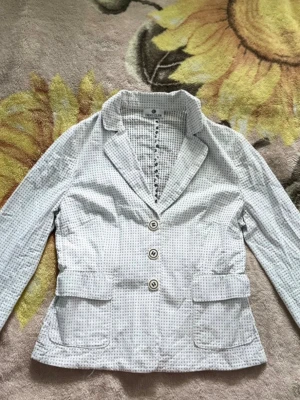 Vit rutig kavaj från Deni Cler - Snygg vit kavaj från Deni Cler med svart rutigt mönster och tre dekorativa knappar framtill. Jackan har klassisk krage, två fickor med lock och långärmad design. Perfekt för dig som gillar stilrena och unika detaljer.