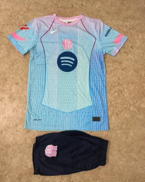 Barça Lamine Yamal fotbollströja set - Säljer ett FC Barcelona matchställ med Lamine Yamal #10. Tröjan är ljusblå med rosa och marinblå detaljer, klubbmärke och sponsortryck. Shortsen är mörkblå med klubbmärke. Perfekt för dig som älskar fotboll och vill sticka ut på planen.