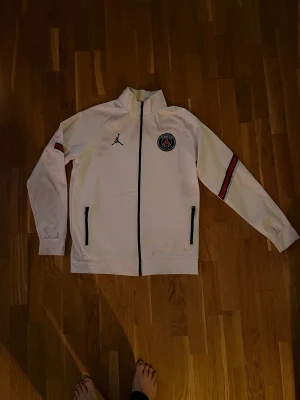 Vit PSG x Jordan ziptröja - Säljer en vit ziptröja från PSG x Jordan med svarta detaljer och dragkedjor. Tröjan har klubbmärke på bröstet, Jordan-logga och röd-svart rand på ärmen. Perfekt för dig som gillar fotboll och streetstyle.