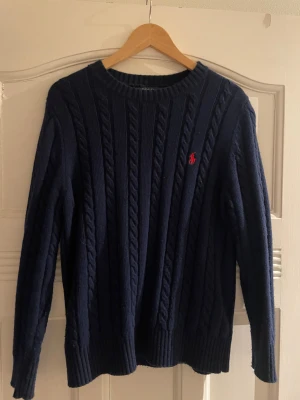 Mörkblå stickad tröja Polo Ralph Lauren - Kabelstickad mörkblå tröja från Polo Ralph Lauren med rund halsringning och röd broderad logga på bröstet. Klassisk modell med ribbade muddar och lång ärm, perfekt för lager-på-lager under höst och vinter.