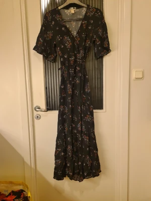 Svart blommig långklänning H&M - Svart långklänning från H&M med v-ringning och korta puffärmar. Klänningen har ett fint blommigt mönster i lila, orange och vitt samt ett smalt knytband i midjan. Materialet är lätt och luftigt, perfekt för varmare dagar.