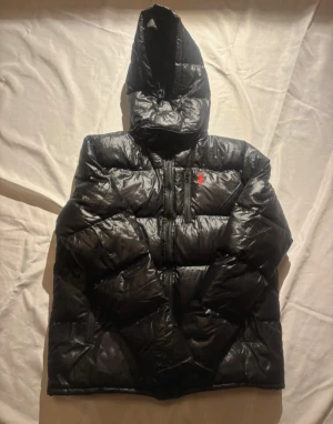 Svart dunjacka från Polo Ralph Lauren - Svart dunjacka från Polo Ralph Lauren med röd broderad logga på bröstet. Jackan har huva, dragkedja framtill och quiltad design. Perfekt för kalla dagar med sin puffiga look och stilrena vibe.