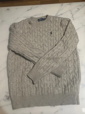 Grå kabelstickad tröja Polo Ralph Lauren - Pris kan pratas om.Snygg grå kabelstickad tröja från Polo Ralph Lauren med klassisk rund hals och broderad logga på bröstet. Tröjan har långa ärmar och ribbade muddar vid ärmslut och nederkant. Perfekt för dig som gillar stilren och tidlös design.