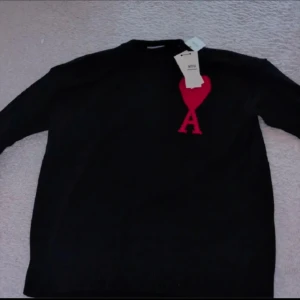 Svart stickad tröja från Ami Paris - Svart stickad tröja från Ami Paris med lång ärm och ikonisk röd hjärtlogga med bokstaven A på bröstet. Klassisk rund halsringning och mjuk ullkvalitet. Perfekt för dig som gillar stilrena plagg med en twist. ----Bilden är tagen Innan användning så Det finns ingen TAG kvar