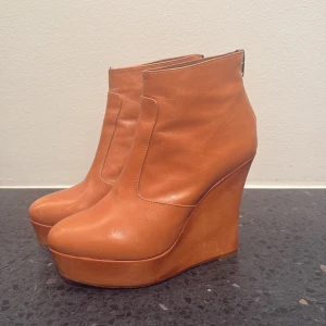 Bruna wedge boots från Novita - Säljer ett par snygga bruna boots från Novita med hög kilklack och plattform. Skorna är tillverkade i skinn och har rund tå samt dragkedja baktill för enkel på- och avtagning. Perfekta för dig som vill ha extra höjd och stil. Ny pris är 2200 jag vill gärna ha 600 men kan diskuteras 