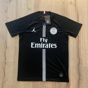 Svart PSG fotbollströja Jordan - Snygg svart PSG fotbollströja från samarbetet med Jordan. Tröjan har korta ärmar, vit PSG-logga på bröstet, Jumpman-logga och Fly Emirates-tryck. Tunn och lätt polyester med en smal rand i mitten. Perfekt för dig som älskar fotboll och streetstyle.