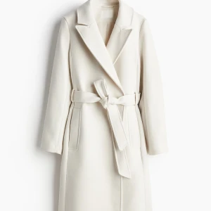 Vit kappa med bälte och slag H&M  - SLUTSÅLD!Stilren och lång vit kappa med klassiska slag och knytskärp i midjan. Kappan har raka linjer, diskreta fickor och är helfodrad. Perfekt för dig som vill ha en clean och elegant look under kyliga dagar.