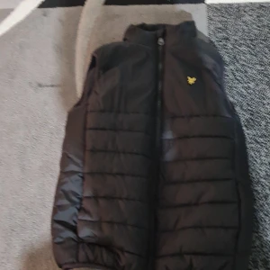 Svart dunväst från Lyle & Scott Junior - Svart dunväst från Lyle & Scott Junior med gul logga på bröstet. Västen har quiltad design, hög krage och dragkedja framtill. Perfekt för lager-på-lager och enkel att matcha med hoodies eller tröjor.