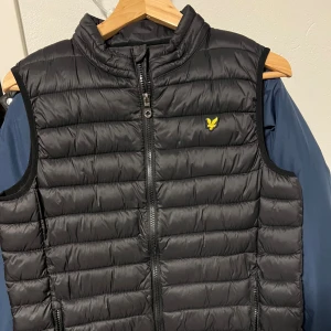 Svart dunväst från Lyle & Scott - Svart quiltad dunväst från Lyle & Scott med gul logotyp på bröstet. Västen har hög krage, dragkedja framtill och två fickor med dragkedja. Perfekt för lager-på-lager och enkel att matcha med olika outfits.