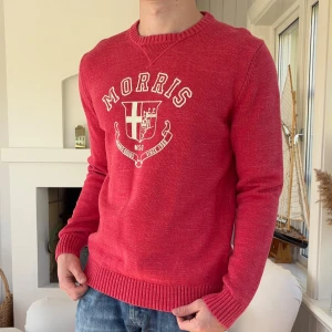 Röd stickad tröja från Morris - Röd stickad tröja från Morris med broderad logga och emblem framtill. Tröjan har rund halsringning, ribbade muddar och är långärmad. Perfekt för dig som gillar klassisk och stilren design med en sportig touch.
