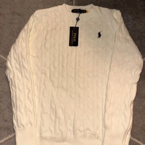Vit kabelstickad tröja Polo Ralph Lauren - Kabelstickad vit tröja från Polo Ralph Lauren med klassisk rund hals och broderad logga på bröstet. Tröjan har lång ärm och ribbade muddar för en snygg passform. Perfekt för dig som gillar stilren och tidlös design.