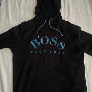 Svart hoodie från Hugo Boss med blå logga - Svart hoodie från Hugo Boss med stor blå BOSS-logga framtill och matchande blå snören i huvan. Klassisk känguruficka och mjukt material som känns skönt mot huden. Perfekt för dig som gillar stilrena streetwear-plagg med tydlig branding.