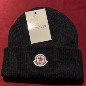 Svart Moncler mössa i ull - Svart ribbstickad mössa från Moncler med klassisk uppvikt kant och broderad logotyp framtill. Tillverkad i mjuk ull som håller dig varm under kyliga dagar. Perfekt accessoar för dig som vill ha en stilren och exklusiv look.