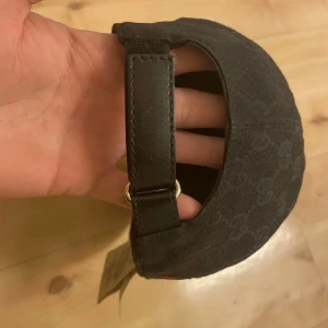 Helt äkta  - Säljer en svart keps från Gucci med klassiskt GG-monogram över hela kepsen. Kepsen har justerbart spänne bak i skinn och ikoniska gröna och röda ränder på sidan. Tillverkad i bomull med detaljer i skinn, perfekt för dig som gillar exklusiva accessoarer.