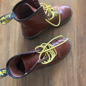 Dr. Martens - Säljer ett par klassiska Dr. Martens boots i oxblod skinn med gula snören och svart kant. Grov sula med tydligt mönster och gul söm runt sulan. Ikonisk Dr. Martens-hälflik och robust känsla. Perfekta för dig som vill sticka ut med en tidlös stil.