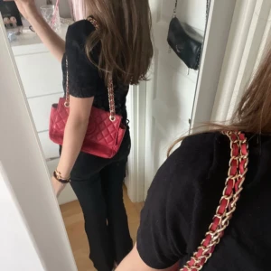 Röd quiltad axelväska från Guess - Snygg röd axelväska från Guess med quiltad design och guldfärgad kedja med röda skinninslag. Väskan har ett stort GG-logo framtill och stängs med magnetlås. Perfekt storlek för det viktigaste och ger en lyxig vibe till din outfit. Den har några fläckar och slitage på insidan och lite sönder på axelbandet. 