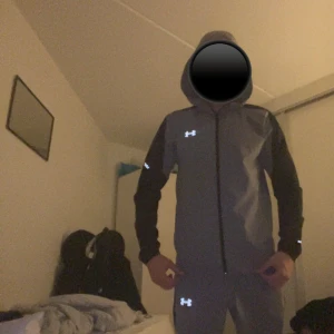 Grå vindset från Under Armour - Snygg grå vindjacka från Under Armour med svarta ärmar och huva. Jackan har dragkedja framtill och reflekterande logga på bröstet. Köpt för 229kr från under armours hemsida 