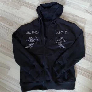 Svart rhinestone hoodie från Blinglucid - Svart hoodie från Blinglucid med huva och dragkedja. Framsidan har coola strasstenar som bildar texten 'BLING LUCID' och två cupider med pilbåge. Perfekt för dig som gillar statement-plagg och streetstyle.
