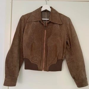 Brun mocka bomberjacka med dragkedja - Snygg brun bomberjacka i mocka med bred krage och ribbade muddar. Jackan har två fickor framtill och stängs med en kraftig dragkedja. Märken bak på jackan och vid kragen, inte testat att kemtvätta.