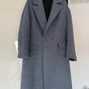 Grå ullkappa med guldknappar - Säljer en lång, grå kappa i ull med snygga guldknappar framtill och på ärmarna. Klassisk krage, dubbla fickor och rak passform. Perfekt för dig som vill ha en stilren och tidlös look under kyliga dagar.