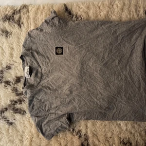 Grå t-shirt från Stone Island - Snygg grå t-shirt från Stone Island med klassisk logga på bröstet. T-shirten är i mjuk bomull och har en enkel, stilren design med rund hals och korta ärmar. Perfekt för en clean och avslappnad look. Priset ej hugget i sten 