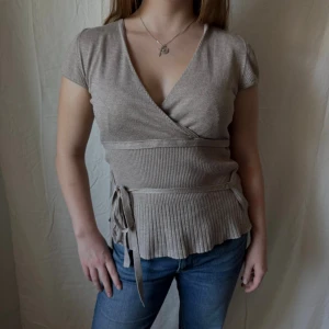 Beige omlottopp med knyt - Snygg beige omlottopp med v-ringning och korta ärmar. Toppen har ribbad midja och nederkant samt knytband i sidan. Perfekt att både till vardags och finare tillfällen. Storlek L men passar S/M, modellen på bilden har vanligtvis S.