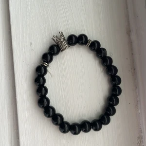 Svart pärlarmband med krona - Snyggt svart pärlarmband med en silverfärgad krona som detalj. Armbandet består av glansiga, runda pärlor och har små silverringar som extra touch. Perfekt för dig som vill sticka ut med en cool accessoar.