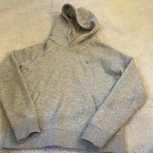 Grå hoodie från GANT med broderad logo - Snygg ljusgrå hoodie från GANT med broderad logo och sköld på bröstet. Tröjan har huva, känguruficka och ribbade muddar vid ärmslut och nederkant. Perfekt för chill dagar och enkel att matcha med jeans eller joggers.