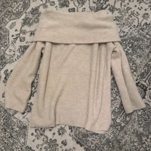 Beige offshoulder stickad tröja H&M - Superfin beige stickad tröja från H&M med bred offshoulder-krage. Tröjan har ribbad struktur och långa ärmar, perfekt för lager-på-lager under hösten och vintern. Snygg att styla med jeans eller kjol för en chill men trendig look.