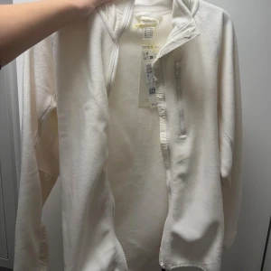 Vit fleecejacka från H&M Move - Snygg vit fleecejacka från H&M Move med dragkedja framtill och en bröstficka med zip. Jackan har långärmade ärmar och paneler i ett annat tyg på underarmen för extra stil. Perfekt för dig som gillar sportig och clean look.