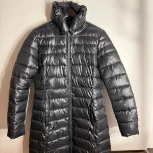 Svart lång dunjacka från Yest - Säljer en svart, lång dunjacka från Yest med hög krage och horisontella quiltade paneler. Jackan har dragkedja framtill och två sidofickor. Perfekt för kalla dagar med sin snygga, glansiga finish och klassiska look.