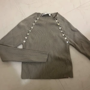 Ribbad tröja med knappar Zara - Säljer en ribbad tröja från Zara med långa ärmar och snygga vita knappar längs axlarna. Superenkel att styla och ett perfekt plagg i garderoben! 