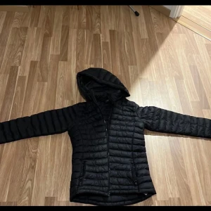 Svart pufferjacka från Everest - Svart pufferjacka från Everest med huva och dragkedja framtill. Jackan har quiltad design, två fickor med dragkedja och lång ärm. Perfekt för kalla dagar och har en stilren look som funkar till de flesta outfits.