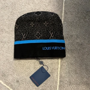  svart mössa med blå kant - Snygg svart mössa från  med klassiskt monogrammönster i grått och en blå rand längst ner. Märkets logga är broderad i blått framtill. Perfekt accessoar för att lyfta din vinterstil.