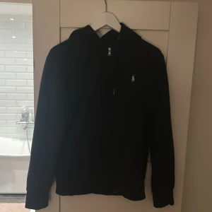 Svart hoodie från Polo Ralph Lauren - Svart hoodie från Polo Ralph Lauren med dragkedja framtill och klassisk logga broderad på bröstet. Tröjan har huva med snörning och långa ärmar. Perfekt för en chill och stilren look.